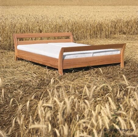 Holzbett mit weißer Matratze steht in einem goldenen Getreidefeld unter freiem Himmel.