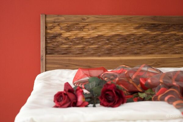 Holzbett mit geschnitztem Kopfteil, roten Rosen und Dekostoff vor roter Wand.