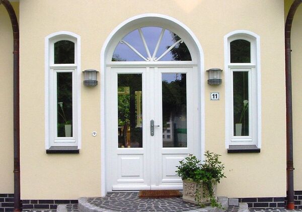 Weiße Haustür mit modernen Fenstern und Pflanzen vor einem gelben Einfamilienhaus.